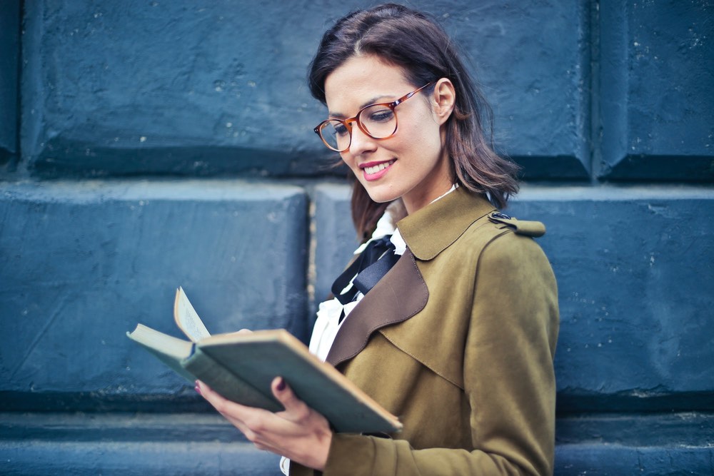 Phot d'une femme de 30 ans, jeune cougar pour un site de rencontre. Elle a des lunettes est vêtue d'un manteau couleur caca d'oie et elle lit un livre en souriant